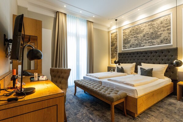 A hotel room with a bed, a desk, a chair, a bench, and a large framed artwork on the wall.