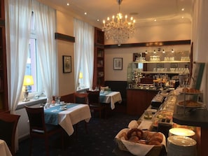 A dining area with tables set for breakfast, a buffet with bread and pastries, and a bar area with glasses and bottles. - Hotel West (Frankfurt)