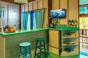 Cozinha compacta com bancada verde, uma pequena geladeira, TV fixada na parede e uma janela com cortinas azuis.
