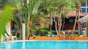 Un espace piscine doté de chaises longues et d’une terrasse en bois, entouré de plantes tropicales et bordé d’un bâtiment au toit rouge.