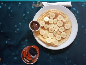 Un plato de tortitas cubiertas con rodajas de plátano y un cuenco pequeño de salsa de chocolate, sobre una mesa con mantel oscuro.
