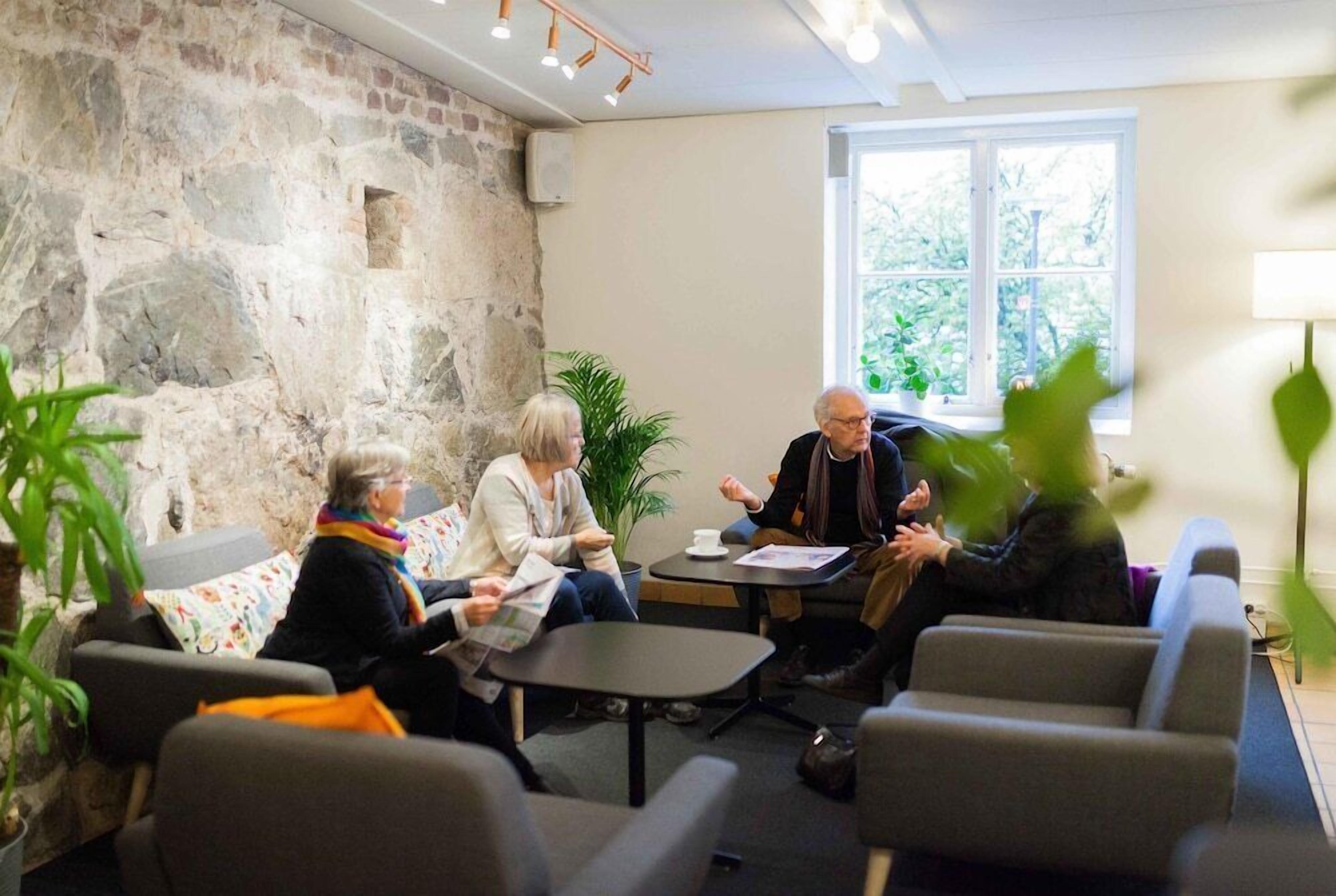 Een groep mensen zit in een kamer met stenen muren, een raam en binnenplanten.