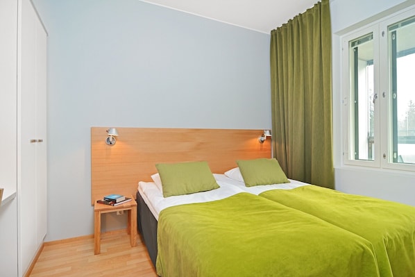 Un dormitorio con cabecera de madera, dos almohadas, colcha verde, mesita de noche con lámpara y ventana con cortinas.