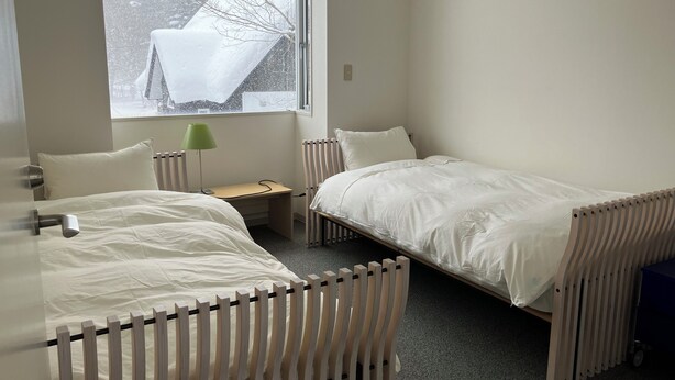 Ein Zimmer mit zwei Betten, einem Schreibtisch, einer Lampe und einem Fenster mit Blick auf Bäume und ein verschneites Dach.