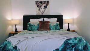 Un dormitorio con una cama, dos mesitas de noche, una lámpara de pared y una obra de arte enmarcada sobre la cama.