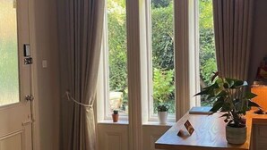 Una habitación con piso de madera, una puerta blanca, una ventana con cortinas, una araña y una pequeña mesa con una planta.