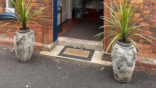Un área de recepción con un letrero, dos cestas de flores colgantes y dos plantas en macetas.