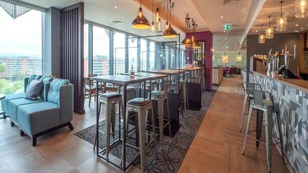 Un espace bar moderne avec des tables hautes et des tabourets, de grandes fenêtres offrant une vue sur la ville, ainsi qu’un mélange de tabourets de bar et d’un canapé.