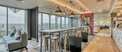 Un hall d’hôtel moderne doté d’un bar, de coins salon et de grandes fenêtres offrant une vue sur la ville.