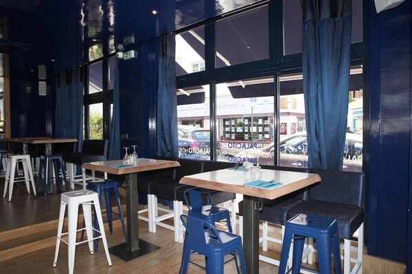 A restaurant interior with tables and chairs, large windows, and a view of the street outside. - Hôtel Palacito (Biarritz)