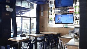 A modern bar area with wooden tables and chairs, a large TV screen, and a variety of bottles on shelves. - Hôtel Palacito (Biarritz)