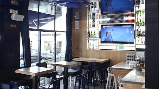 A modern bar area with wooden tables and chairs, a large TV screen, and a variety of bottles on shelves. - Hôtel Palacito (Biarritz)