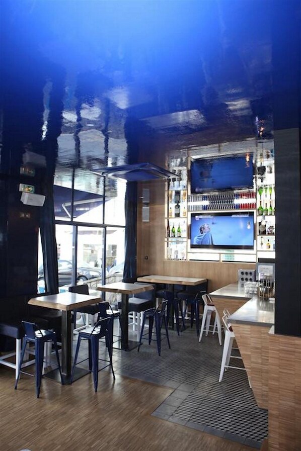 A modern bar area with wooden tables and chairs, a large TV screen, and a variety of bottles on shelves. - Hôtel Palacito (Biarritz)