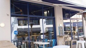 An outdoor seating area with tables and chairs, a stone wall, and a building with large windows and a blue awning. - Hôtel Palacito (Biarritz)