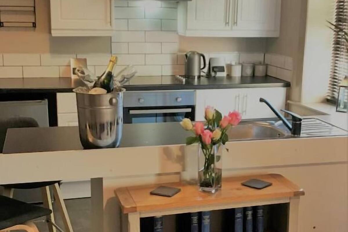Una cocina moderna con armarios blancos, una campana extractora de acero inoxidable, una cafetera y una pequeña zona de bar con estantería.