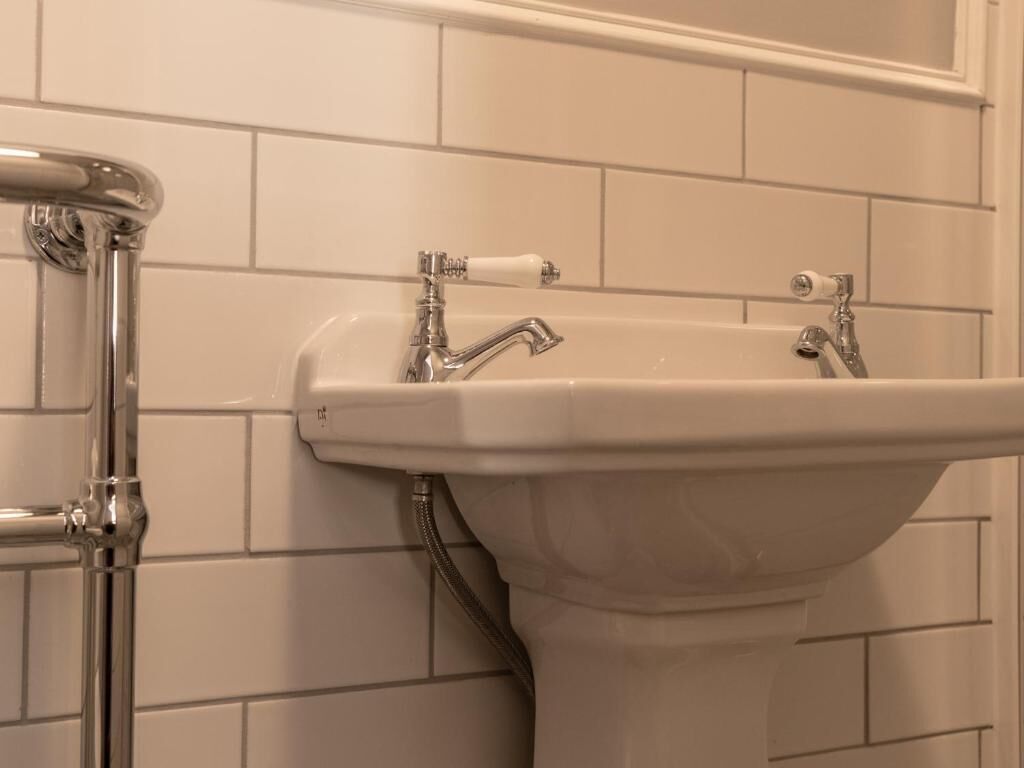 Une salle de bain avec des murs recouverts de carreaux blancs, un lavabo blanc doté de deux robinets et un tuyau chromé.