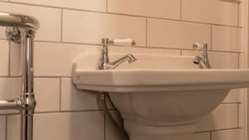 A bathroom with white tiled walls, a white sink with two faucets, and a chrome pipe.