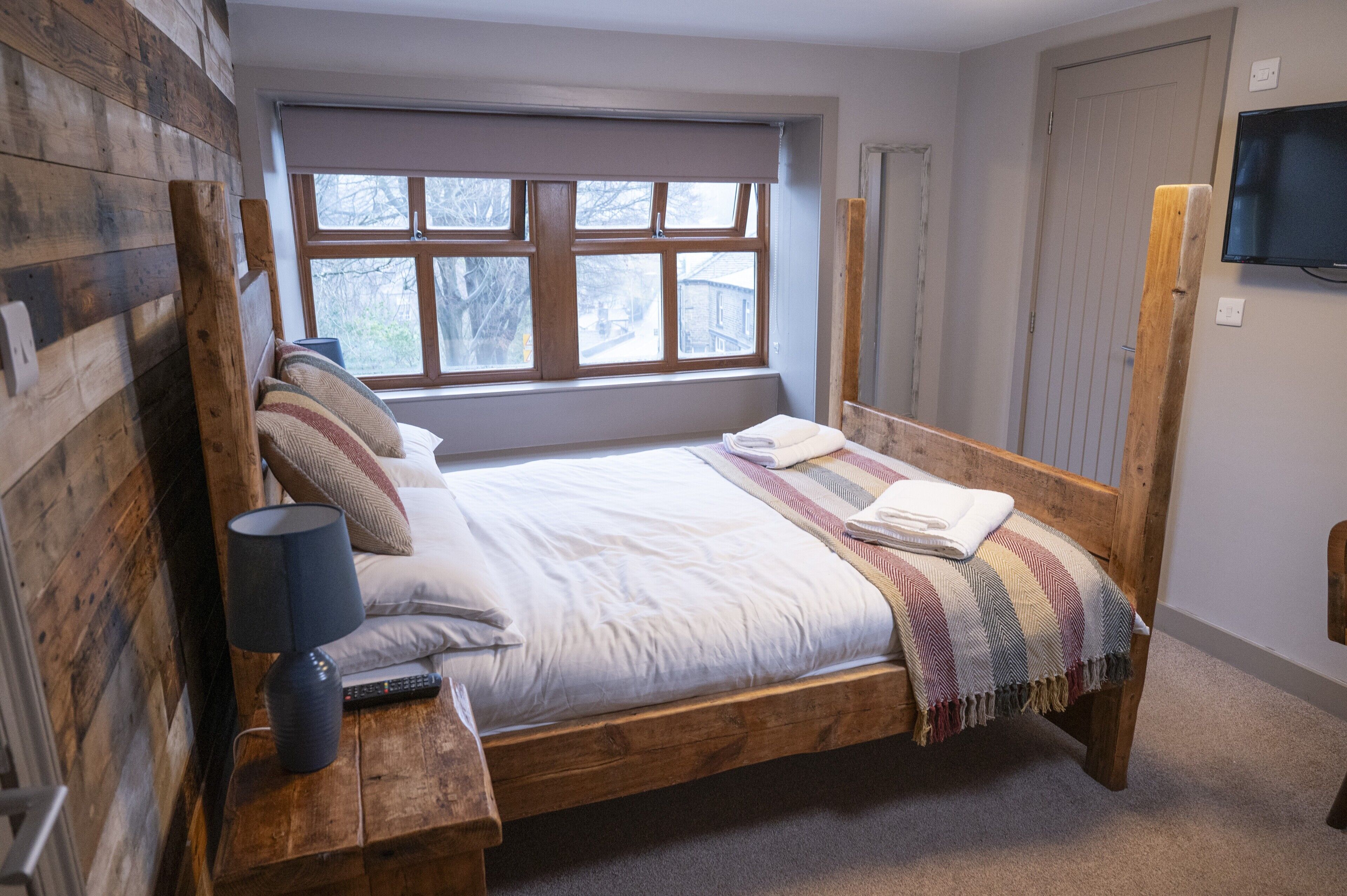 A wooden bed with a window above, a TV on the wall, and a lamp on the nightstand.