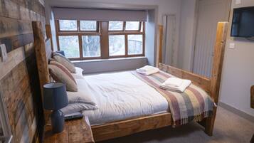 A wooden bed with a window above, a TV on the wall, and a lamp on the nightstand.