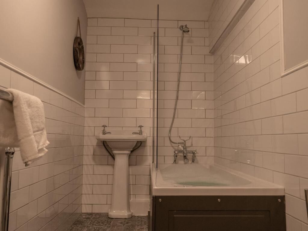 Un baño con paredes de azulejos blancos, un lavamanos pedestal, una bañera y una ducha con mampara de vidrio.