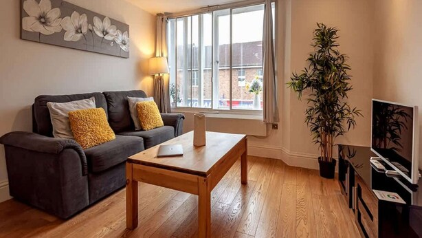 Ein Wohnzimmer mit einer Couch, einem Couchtisch, einer Lampe, einem Fenster mit Vorhängen und einer Topfpflanze.
