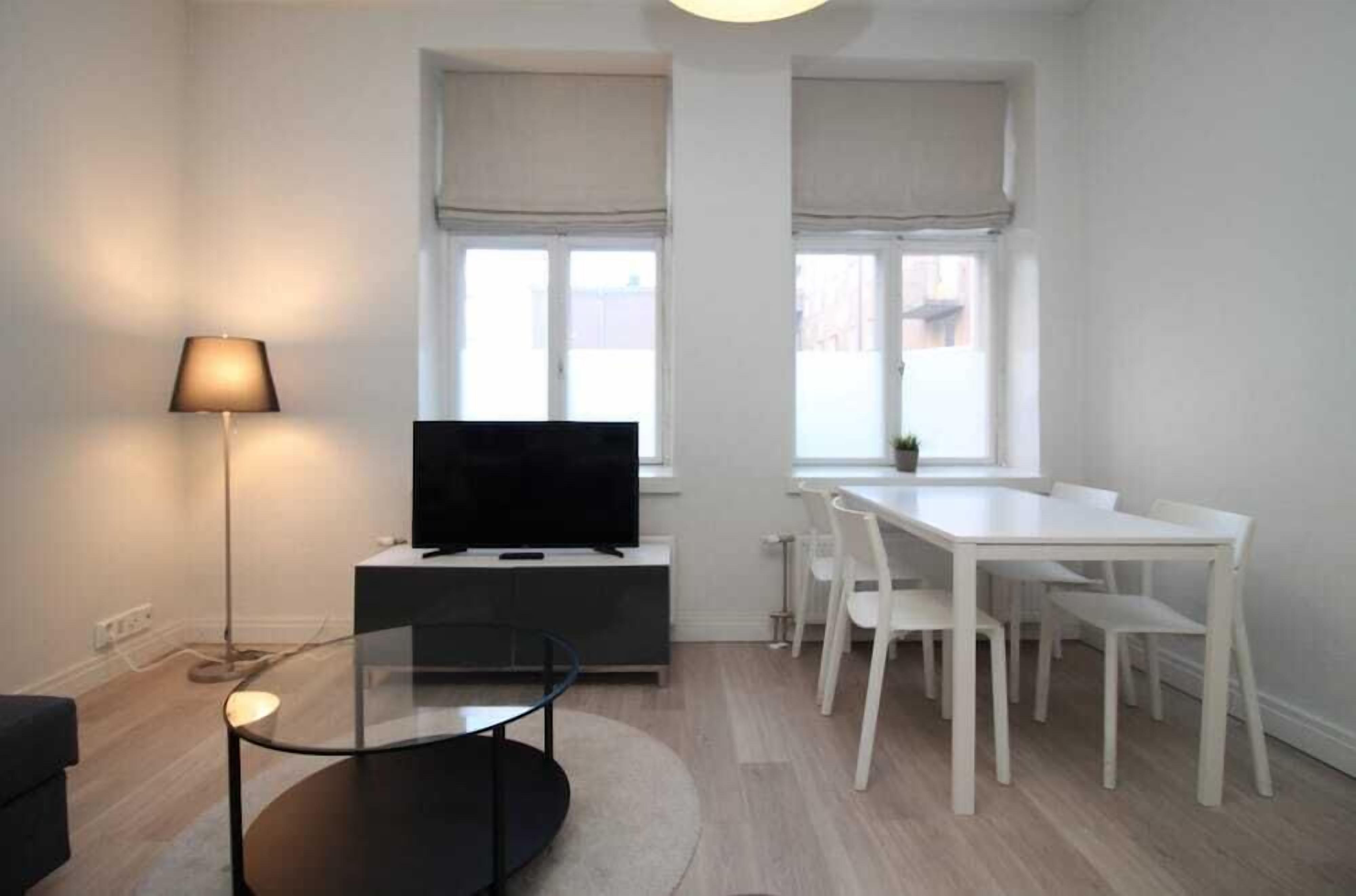 Ein modernes Wohnzimmer mit Flachbildfernseher, einem Glastisch, einem weißen Esstisch mit Stühlen und einer Stehlampe.