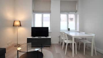 A modern living room with a flat-screen TV, a glass coffee table, a white dining table with chairs, and a floor lamp.