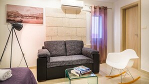 A living room with a sofa, a white chair, a glass coffee table with books, an air conditioner, and a painting on the wall. - Split Oasis Apartments (Split)