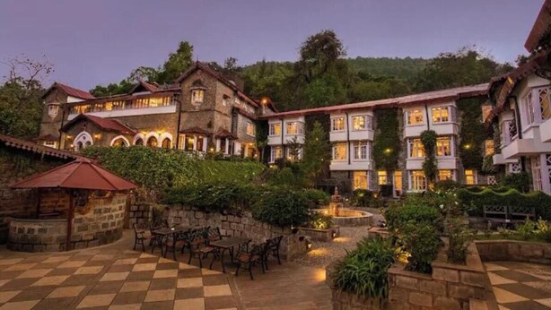 Un patio con mesas y sillas de piedra, rodeado de edificios con ventanas arqueadas y una fuente.