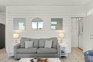 Ein modernes Wohnzimmer mit einem grauen Sofa, zwei Beistelltischen mit Lampen, einem runden Spiegel und gerahmten Bildern an der Wand.