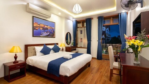 A hotel room with a wooden floor, a bed with blue and white bedding, a desk with a chair, a desk lamp, a wall-mounted fan, and a painting of a coastal scene. - The Linh Hoi An (Hoi An)