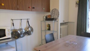 Uma cozinha compacta com micro-ondas, panelas penduradas, uma prateleira com garrafas e uma mesa de jantar com cadeiras.