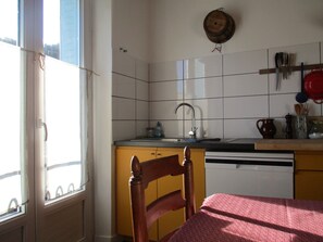 Een keuken met een raam, een spoelbak, een houten stoel en een tafel met een tafelkleed met dessin.