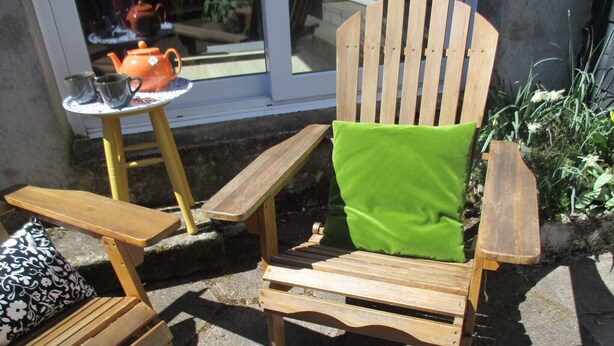 Uma cadeira Adirondack de madeira com almofada verde, uma mesinha com uma chaleira e uma janela com vista para plantas.