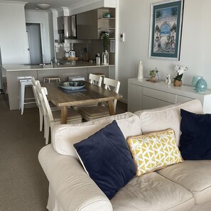 Uma sala de estar moderna com um sofá bege, uma mesa de jantar de madeira e uma cozinha ao fundo.
