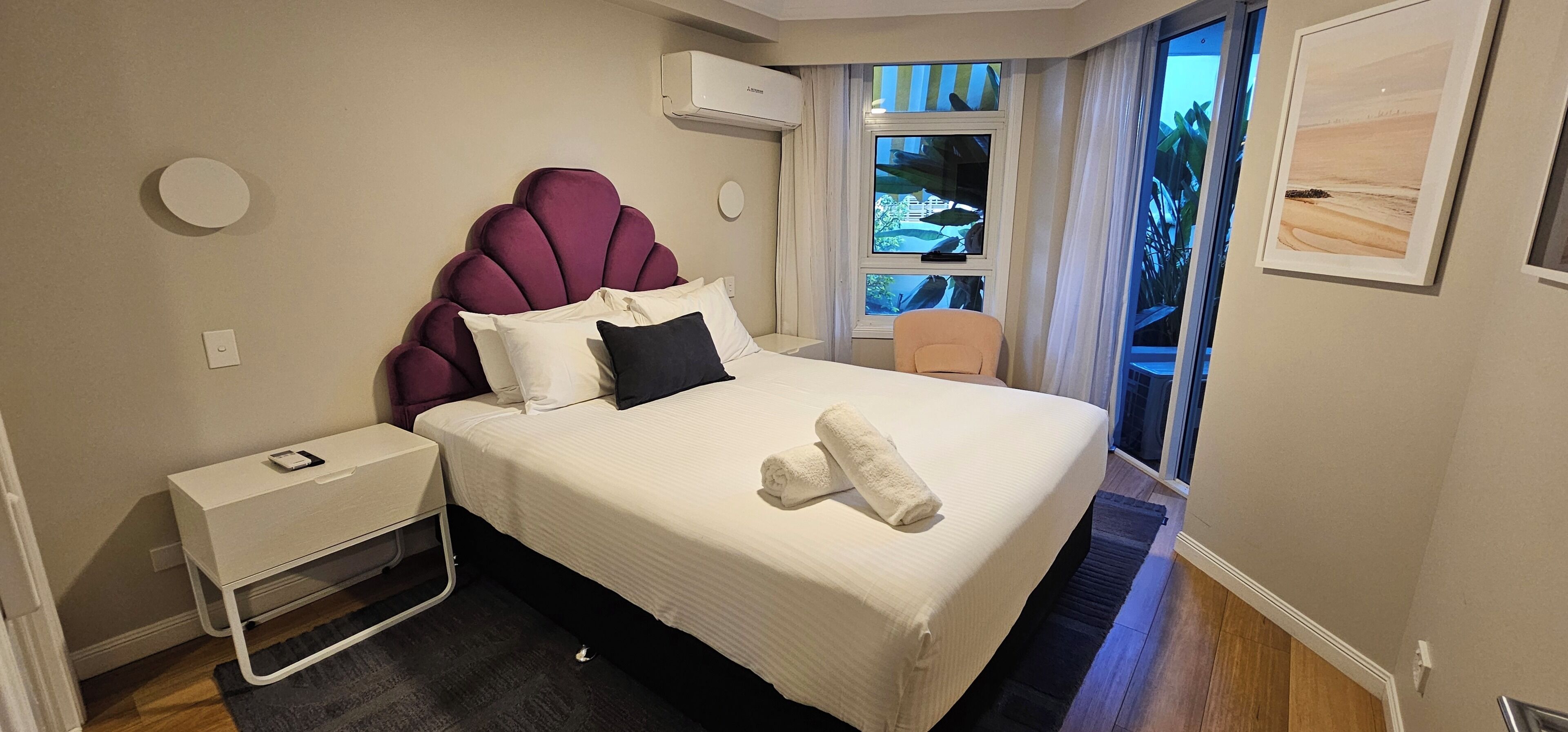 A hotel room with a bed, a nightstand, a window with curtains, and a framed picture on the wall.