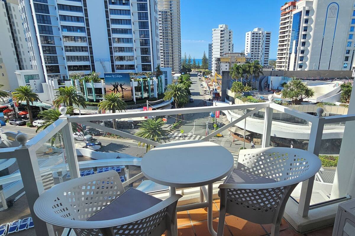 Un balcón con dos mesas y sillas blancas, con vista a un paisaje urbano con edificios altos y palmeras.