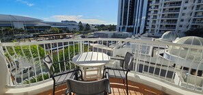 A balcony with a table and chairs, a view of a cityscape including a tall building and a stadium, and a fenced area with plants. - Broadbeach Holiday Apartments (Gold Coast)