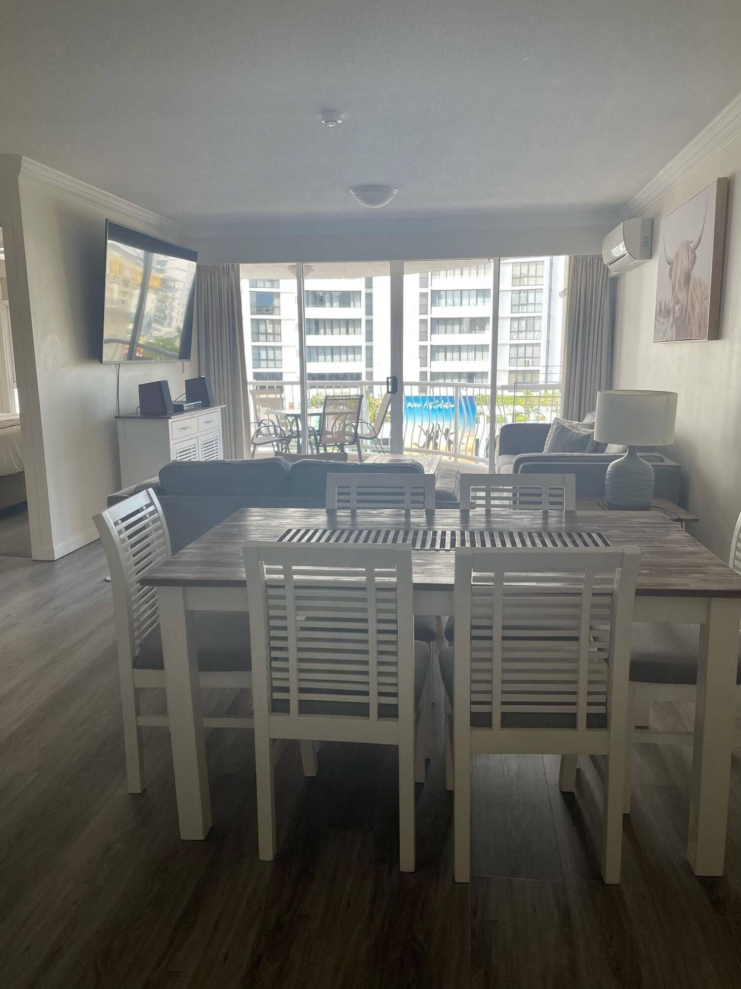 A modern living room with a dining area, large windows, a flat-screen TV, and a view of buildings.
