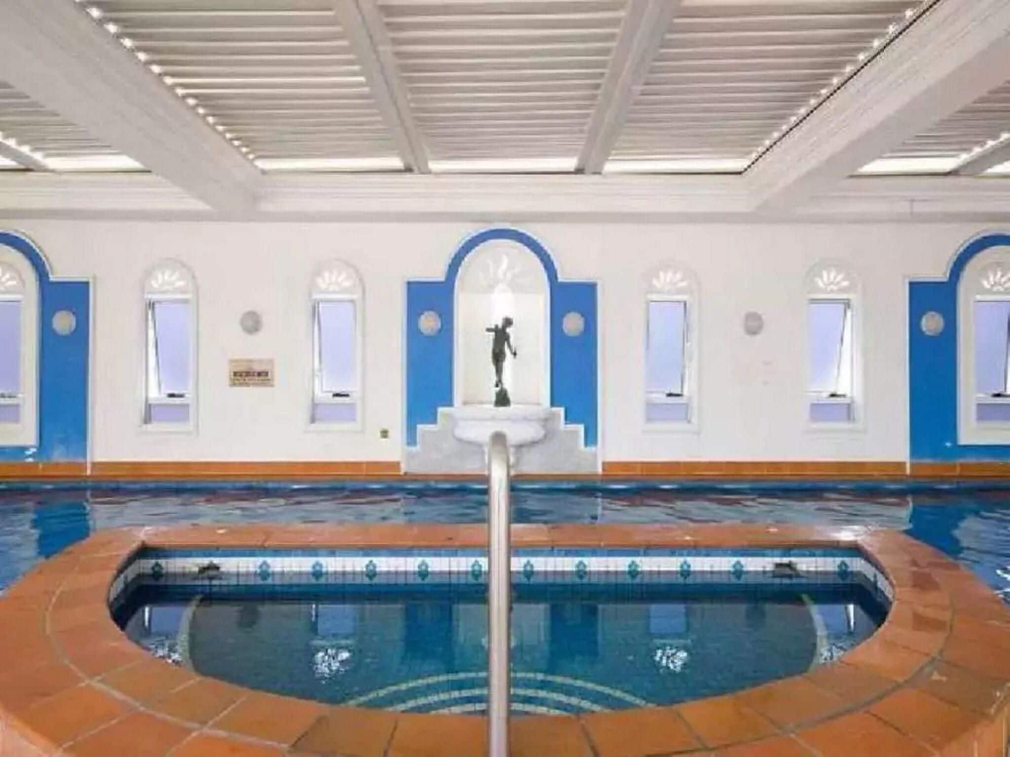 An indoor swimming pool with a tiled edge, a central fountain, and arched windows along the walls.