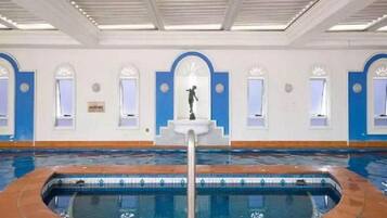 An indoor swimming pool with a tiled edge, a central fountain, and arched windows along the walls.