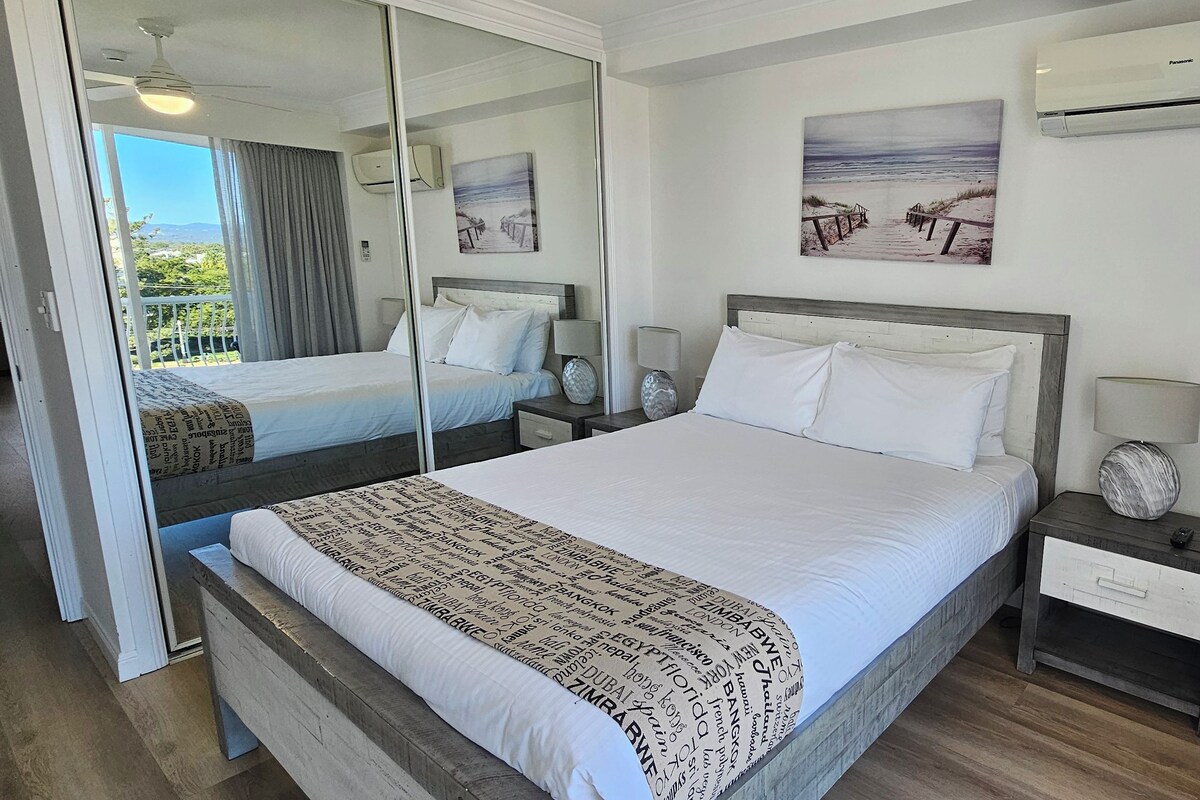 Habitación de hotel con una cama grande, dos mesitas de noche, un espejo y vistas al exterior.