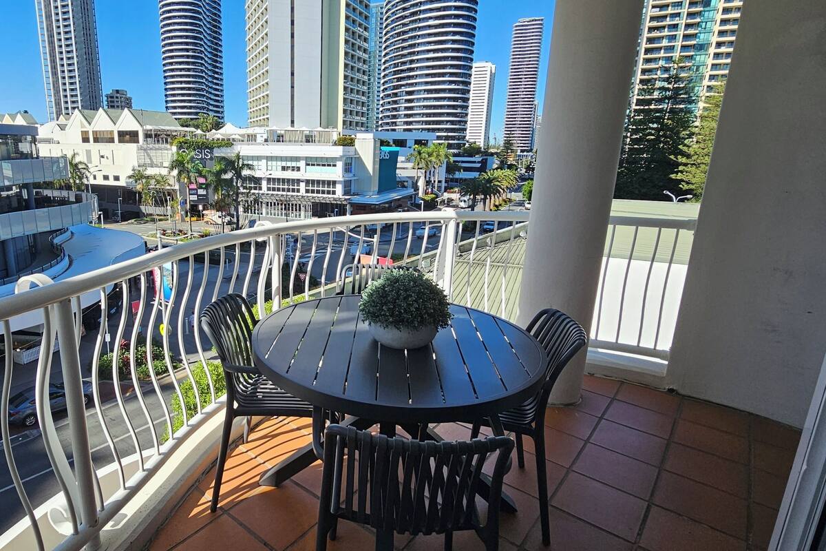 Un balcón con una mesa redonda y sillas, con vistas a un paisaje urbano de edificios modernos y cielo despejado.