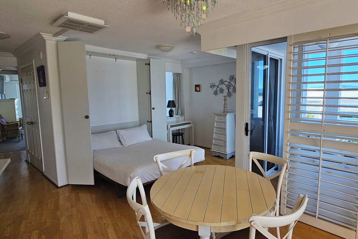A room with a bed, a round wooden table, white chairs, a dresser, and a window with blinds.