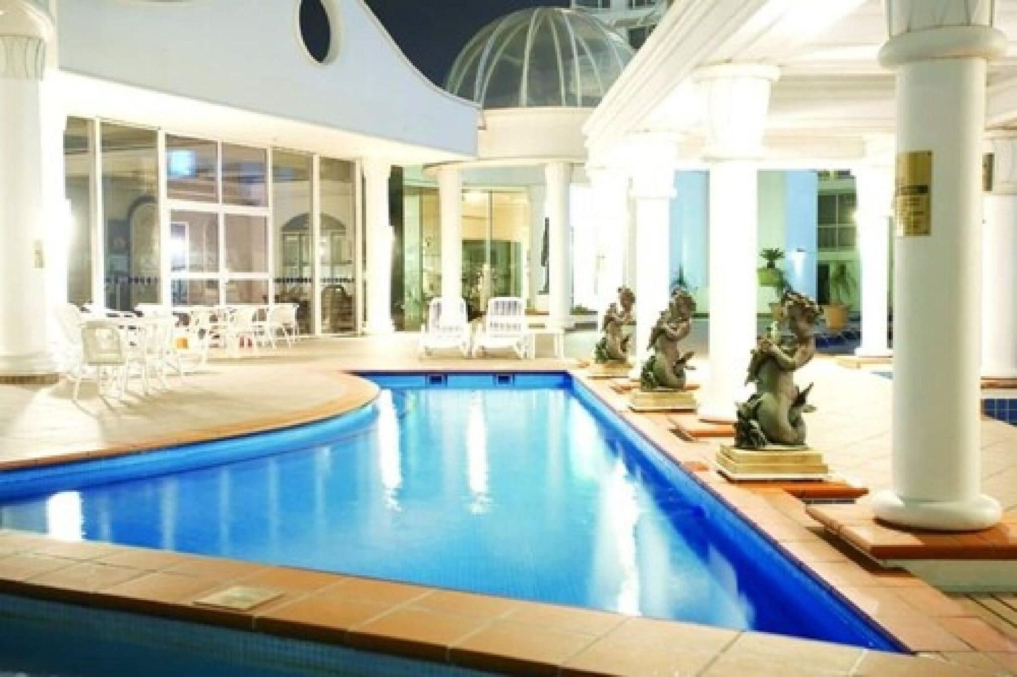 An indoor swimming pool with white columns, a seating area with chairs, and decorative statues.