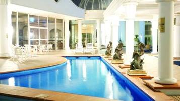 An indoor swimming pool with white columns, a seating area with chairs, and decorative statues.