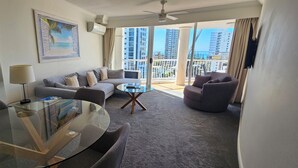 Una sala de estar moderna con una mesa de vidrio, un sofá gris y sillones, con un ventanal que ofrece vistas a rascacielos y al mar.