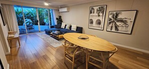 A modern living room with a sofa, coffee table, and a dining area with a wooden table and chairs. There are large windows with a view of greenery outside. - Broadbeach Holiday Apartments (Gold Coast)