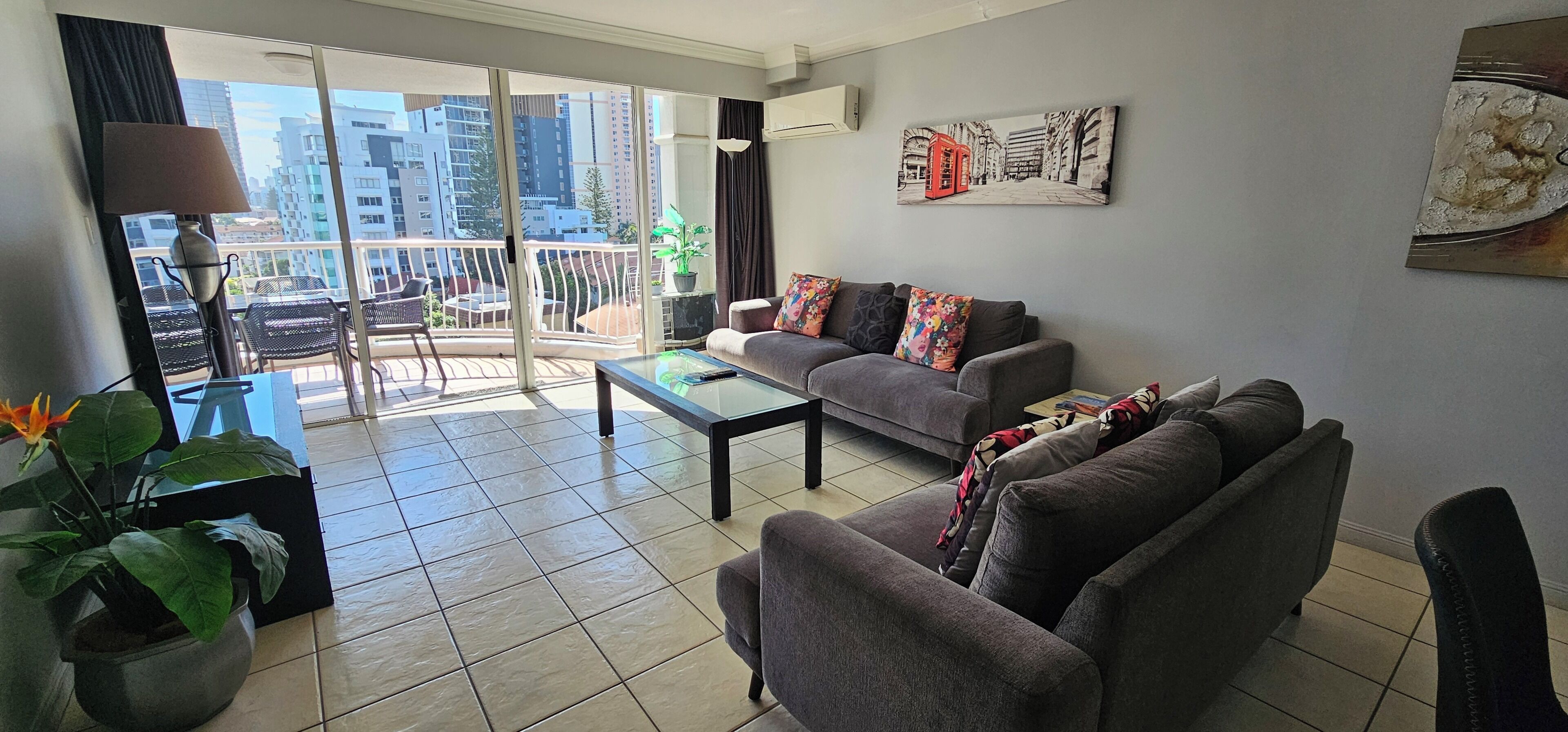 A modern living room with a grey sofa, a glass coffee table, and a balcony with a view of tall buildings.