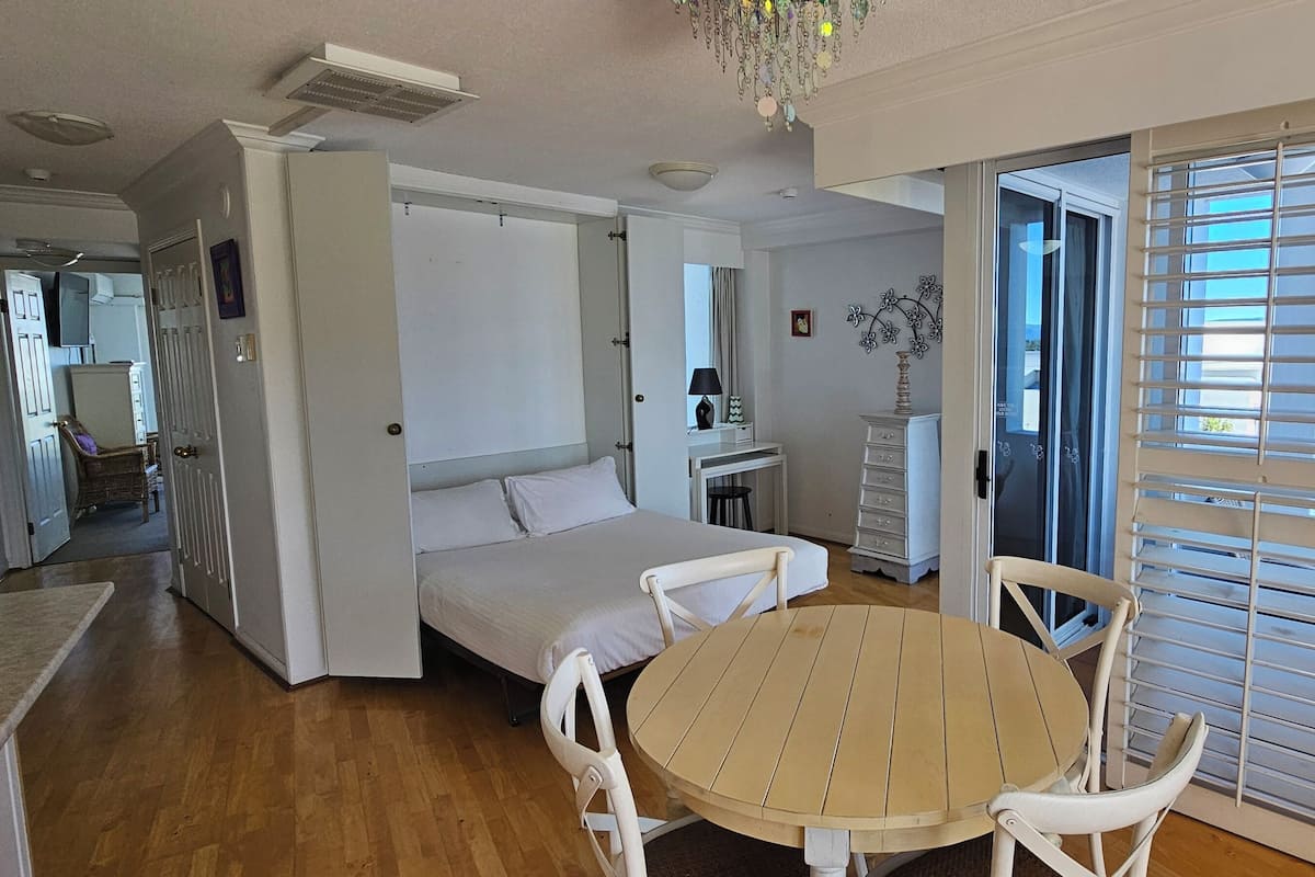 Una habitación con una cama, una mesa redonda de madera, sillas blancas, un aparador y una ventana con persianas.